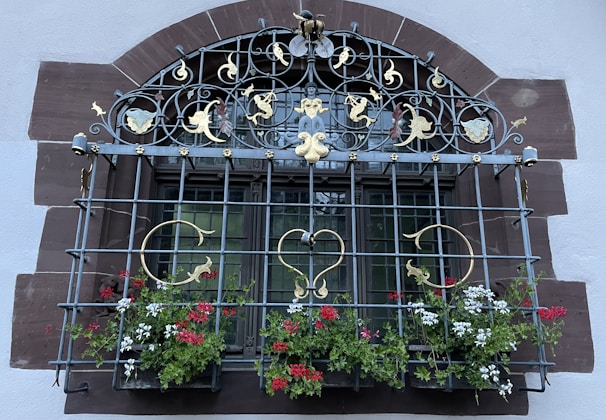 An ornate window with intricate metalwork featuring gold and black elements, including crescents, hearts, and decorative flourishes. The window is adorned with red and white flowers in a planter at the bottom. The frame is set against a stone archway with a mixture of brown and white colors.