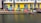 Rows of colorful kayaks and paddleboards are stacked against a yellow and gray building with large windows. The building is situated next to a body of water, likely a canal or river. Some equipment is also placed around, and there is a small dock area.