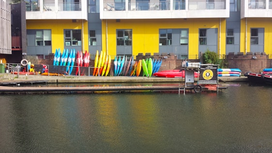 Rows of colorful kayaks and paddleboards are stacked against a yellow and gray building with large windows. The building is situated next to a body of water, likely a canal or river. Some equipment is also placed around, and there is a small dock area.