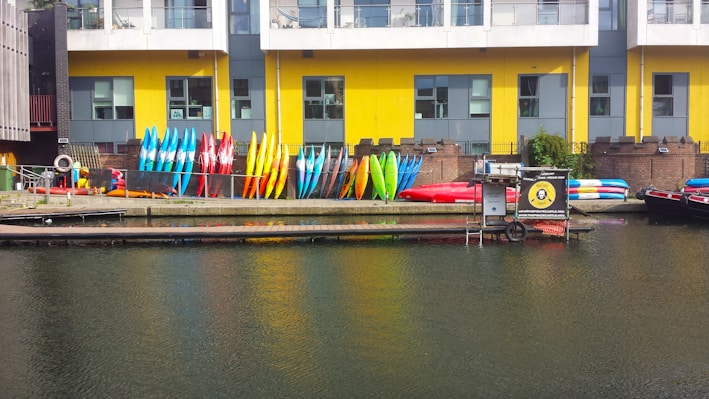 Rows of colorful kayaks and paddleboards are stacked against a yellow and gray building with large windows. The building is situated next to a body of water, likely a canal or river. Some equipment is also placed around, and there is a small dock area.