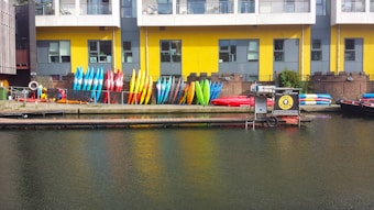 Rows of colorful kayaks and paddleboards are stacked against a yellow and gray building with large windows. The building is situated next to a body of water, likely a canal or river. Some equipment is also placed around, and there is a small dock area.