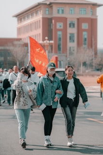 A group of people walking together outdoors, with three women in the foreground smiling and conversing. They are casually dressed and holding papers. Behind them, additional people are visible, carrying a bright orange flag with writing on it. A large building with arched windows and a red brick facade is seen in the background.