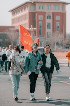 A group of people walking together outdoors, with three women in the foreground smiling and conversing. They are casually dressed and holding papers. Behind them, additional people are visible, carrying a bright orange flag with writing on it. A large building with arched windows and a red brick facade is seen in the background.