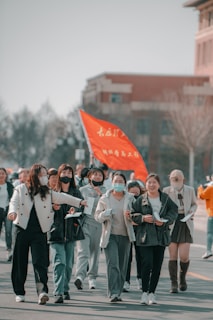 A group of people, mostly women, are walking together in an outdoor setting. They seem to be participating in a gathering or event as some are holding papers and there is a visible red flag with text. The participants are wearing casual or sporty clothing and a few have face masks.