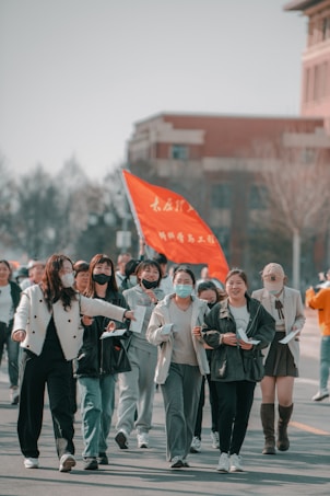 A group of people, mostly women, are walking together in an outdoor setting. They seem to be participating in a gathering or event as some are holding papers and there is a visible red flag with text. The participants are wearing casual or sporty clothing and a few have face masks.