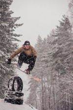 A snowboarder is performing a trick, jumping over a stack of tires in a snowy forest setting. Snow is visibly falling, adding to the wintry atmosphere. The person is dressed in a brown jacket, black pants, and a black helmet.