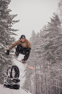 A snowboarder is performing a trick, jumping over a stack of tires in a snowy forest setting. Snow is visibly falling, adding to the wintry atmosphere. The person is dressed in a brown jacket, black pants, and a black helmet.