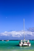 A catamaran is anchored in clear turquoise waters under a deep blue sky. Other small boats are visible in the distance against a backdrop of fluffy white clouds. The scene is tranquil and picturesque, suggesting a tropical setting.