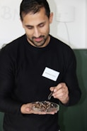 A person is holding a petri dish filled with several insect parts, using tweezers to handle them. They are wearing a black shirt and have a name tag pinned to it. The background is a muted, light-colored wall.