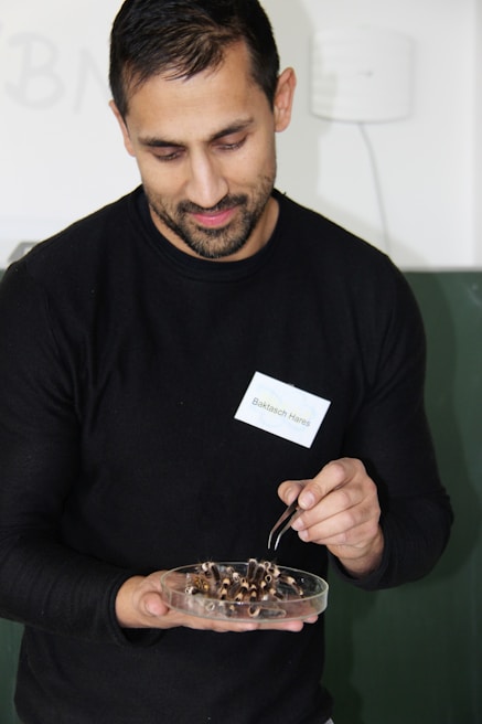 A person is holding a petri dish filled with several insect parts, using tweezers to handle them. They are wearing a black shirt and have a name tag pinned to it. The background is a muted, light-colored wall.