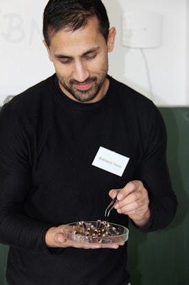 A person is holding a petri dish filled with several insect parts, using tweezers to handle them. They are wearing a black shirt and have a name tag pinned to it. The background is a muted, light-colored wall.