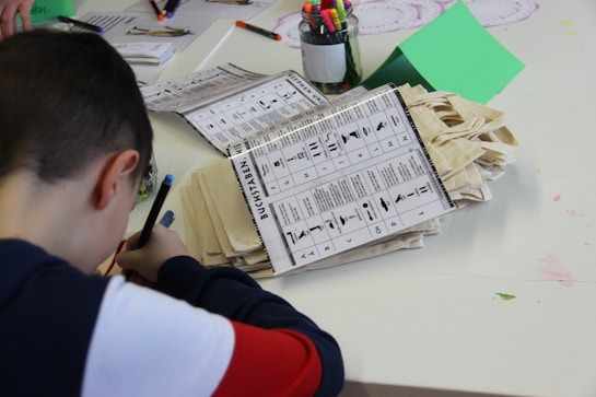A child is drawing or writing at a table covered with various craft materials and sheets of paper. There is a stack of instruction booklets or activity sheets featuring icons and text, likely for a craft or learning activity. Assorted colored markers are held in a clear jar nearby, along with other papers and a green folder.