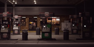 A dimly lit gas station with multiple fuel pumps displaying the Phillips 66 logo. Behind the pumps is a convenience store with illuminated signage and windows showing items for sale inside. There is an ice machine to the right of the convenience store entrance.