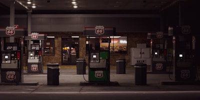 A dimly lit gas station with multiple fuel pumps displaying the Phillips 66 logo. Behind the pumps is a convenience store with illuminated signage and windows showing items for sale inside. There is an ice machine to the right of the convenience store entrance.