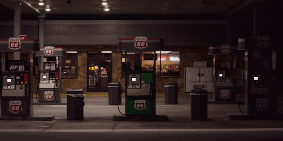 A dimly lit gas station with multiple fuel pumps displaying the Phillips 66 logo. Behind the pumps is a convenience store with illuminated signage and windows showing items for sale inside. There is an ice machine to the right of the convenience store entrance.