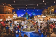 A vibrant, crowded marketplace with numerous stalls selling a variety of goods. The lively atmosphere is accentuated by a large fountain in the foreground and overhead string lights. In the background, there are architectural structures reminiscent of an ancient pyramid, adding a cultural touch to the scene. People are actively engaging, shopping, and exploring the area, with bright lights and colorful decorations enhancing the festive mood.