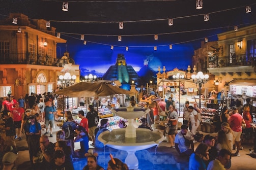 A vibrant, crowded marketplace with numerous stalls selling a variety of goods. The lively atmosphere is accentuated by a large fountain in the foreground and overhead string lights. In the background, there are architectural structures reminiscent of an ancient pyramid, adding a cultural touch to the scene. People are actively engaging, shopping, and exploring the area, with bright lights and colorful decorations enhancing the festive mood.