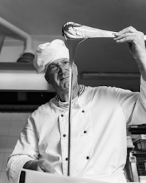 A chef is stretching a piece of dough or candy from a large ladle, with a focused expression. The setting appears to be a kitchen. He is wearing a white chef's uniform and hat.