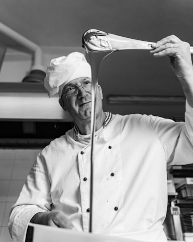 A chef is stretching a piece of dough or candy from a large ladle, with a focused expression. The setting appears to be a kitchen. He is wearing a white chef's uniform and hat.