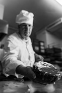 A chef in uniform is holding out a glazed pastry or cake towards the viewer. The setting appears to be a kitchen, with trays and industrial equipment in the background. The focus is on the pastry, capturing its shiny glaze in detail.