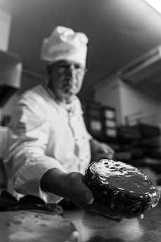 A chef in uniform is holding out a glazed pastry or cake towards the viewer. The setting appears to be a kitchen, with trays and industrial equipment in the background. The focus is on the pastry, capturing its shiny glaze in detail.