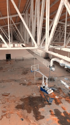 An abandoned indoor space with a basketball hoop and a damaged, stained floor. The ceiling is supported by white metal beams, and there are overhead ducts visible. The area looks neglected with debris scattered around.