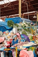 A busy market stall displays a colorful variety of fresh produce, including cauliflower, greens, onions, bananas, and peppers. An elderly woman, wearing a blue apron and headscarf, tends to the stall. A hanging scale and a purple umbrella are also visible. The market is covered by a metal structure.