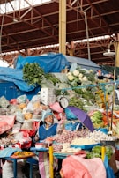 A busy market stall displays a colorful variety of fresh produce, including cauliflower, greens, onions, bananas, and peppers. An elderly woman, wearing a blue apron and headscarf, tends to the stall. A hanging scale and a purple umbrella are also visible. The market is covered by a metal structure.