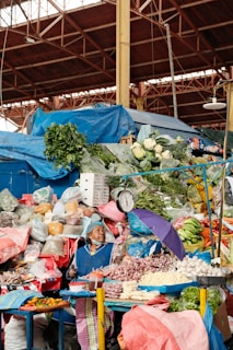 A busy market stall displays a colorful variety of fresh produce, including cauliflower, greens, onions, bananas, and peppers. An elderly woman, wearing a blue apron and headscarf, tends to the stall. A hanging scale and a purple umbrella are also visible. The market is covered by a metal structure.