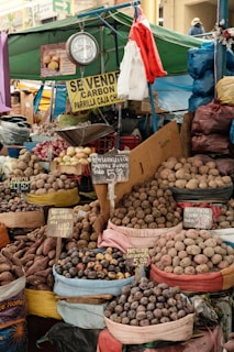 A bustling market stall filled with various types of potatoes and produce, displayed in colorful bags. There are signs in Spanish indicating the names and prices of the items. A traditional weighing scale is prominently hung at the stall. The scene is vibrant with a makeshift canopy of tarpaulins in green and blue, and other market goods in the background.