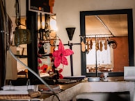 A cozy interior with a collection of hanging accessories, including necklaces and bracelets, displayed on a rack. A vibrant pink decorative flower stands out among the neutral tones. The background features a window with natural light illuminating the space, creating a warm ambiance.