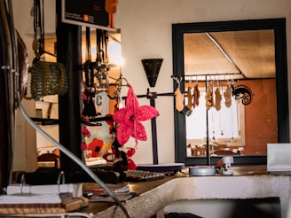 A cozy interior with a collection of hanging accessories, including necklaces and bracelets, displayed on a rack. A vibrant pink decorative flower stands out among the neutral tones. The background features a window with natural light illuminating the space, creating a warm ambiance.