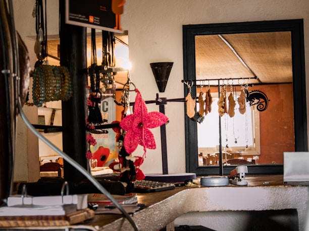 A cozy interior with a collection of hanging accessories, including necklaces and bracelets, displayed on a rack. A vibrant pink decorative flower stands out among the neutral tones. The background features a window with natural light illuminating the space, creating a warm ambiance.
