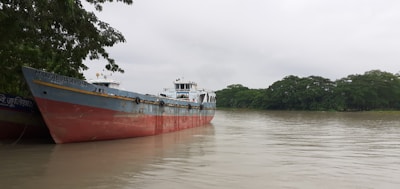 A large cargo ship named M.V. Fabiha Aysha is docked on a calm river, with lush green trees lining the riverbank under a cloudy sky.