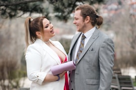 A man and a woman are dressed formally and are smiling at each other. The woman is wearing a white jacket over a pink dress and holding a glittery clutch. The man is in a light gray suit with a patterned tie. They appear to be outdoors with blurred trees in the background.