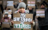 A young person is holding a sign with the message 'No Plastic in Nature'. They are bundled in warm clothing, including a gray beanie, indicating cold weather. In the background, several other people, also wearing winter clothes, hold similar protest signs advocating against plastic.