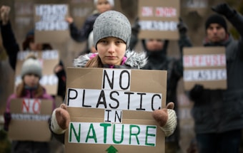 A young person is holding a sign with the message 'No Plastic in Nature'. They are bundled in warm clothing, including a gray beanie, indicating cold weather. In the background, several other people, also wearing winter clothes, hold similar protest signs advocating against plastic.