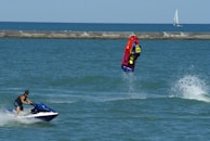 A person riding a jet ski performs a backflip on a vibrant red and yellow watercraft. Another individual on a blue and white jet ski watches nearby. The ocean is calm with a distant sailboat on the horizon and a breakwater separating the water from the sky.