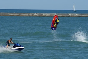 A person riding a jet ski performs a backflip on a vibrant red and yellow watercraft. Another individual on a blue and white jet ski watches nearby. The ocean is calm with a distant sailboat on the horizon and a breakwater separating the water from the sky.