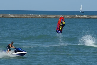 A person riding a jet ski performs a backflip on a vibrant red and yellow watercraft. Another individual on a blue and white jet ski watches nearby. The ocean is calm with a distant sailboat on the horizon and a breakwater separating the water from the sky.