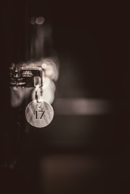 A metallic keychain with the number 17 hangs from a lock in low light, casting soft reflections and shadows. The background is out of focus, creating a bokeh effect with circular light spots.