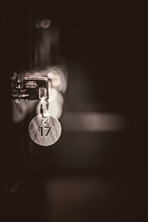 A metallic keychain with the number 17 hangs from a lock in low light, casting soft reflections and shadows. The background is out of focus, creating a bokeh effect with circular light spots.