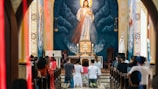 Individuals are kneeling in prayer inside a church. A large depiction of Jesus with a radiant halo is present in the background above the altar. The church interior features ornate designs and warm lighting. The congregation is facing the altar and appears to be engaged in a solemn religious ritual.