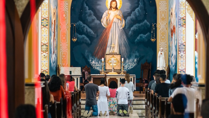 Individuals are kneeling in prayer inside a church. A large depiction of Jesus with a radiant halo is present in the background above the altar. The church interior features ornate designs and warm lighting. The congregation is facing the altar and appears to be engaged in a solemn religious ritual.