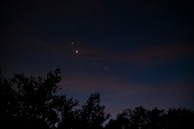 A nighttime sky features two bright celestial objects, likely planets or stars, shining through a dark backdrop. Silhouetted tree tops are visible at the bottom, creating a natural frame for the serene astronomical scene.