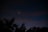 A nighttime sky features two bright celestial objects, likely planets or stars, shining through a dark backdrop. Silhouetted tree tops are visible at the bottom, creating a natural frame for the serene astronomical scene.