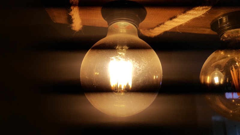 A glowing incandescent light bulb is prominently displayed with its filament brightly shining. The bulb is attached to a fixture and surrounded by a darker, shadowy ambiance with another bulb partially visible.