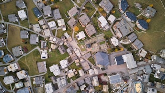 An aerial view of a residential area with numerous buildings, lawns, and roads forming an intricate grid. The buildings are mostly rectangular and vary in size and shape. Green spaces and pathways are interspersed between the structures, giving a balanced look to the urban layout.