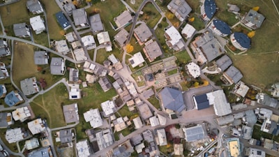 An aerial view of a residential area with numerous buildings, lawns, and roads forming an intricate grid. The buildings are mostly rectangular and vary in size and shape. Green spaces and pathways are interspersed between the structures, giving a balanced look to the urban layout.