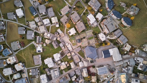 An aerial view of a residential area with numerous buildings, lawns, and roads forming an intricate grid. The buildings are mostly rectangular and vary in size and shape. Green spaces and pathways are interspersed between the structures, giving a balanced look to the urban layout.